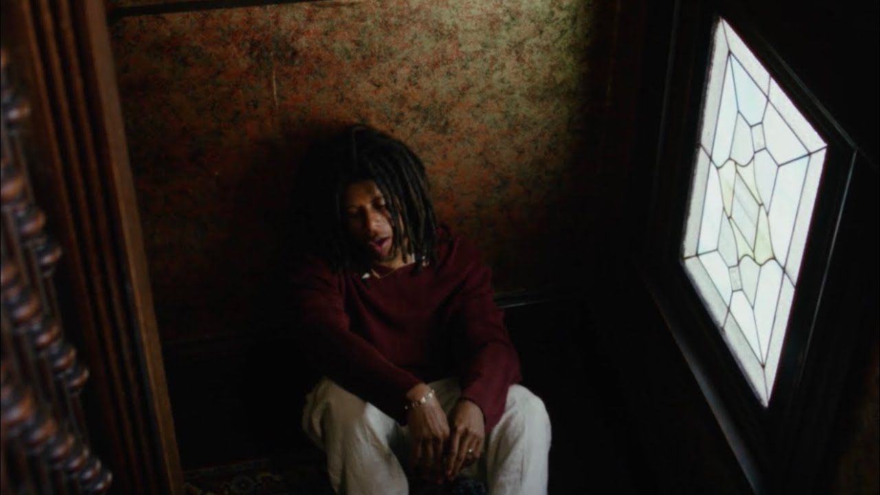Person sitting in a dim corner beside a bright window, head bowed, in a patterned room