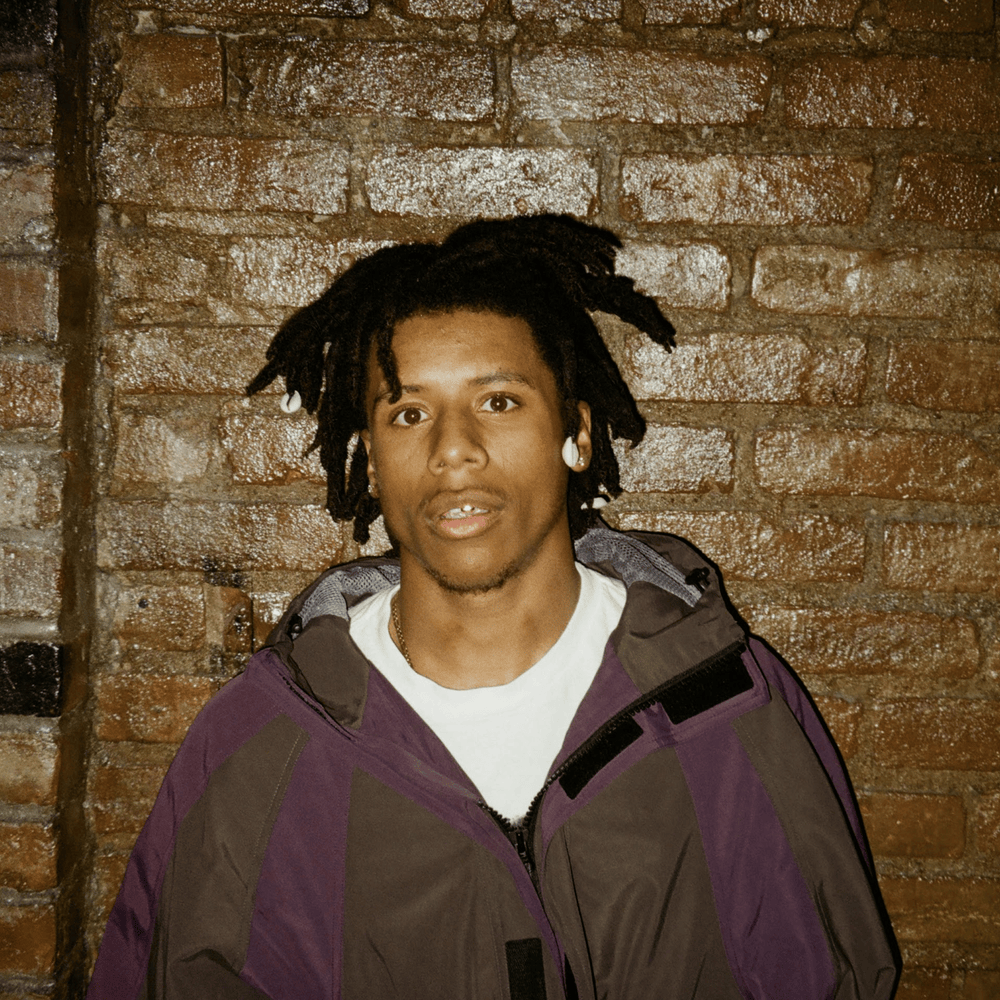 Person in a purple jacket standing against a brick wall, looking at the camera.