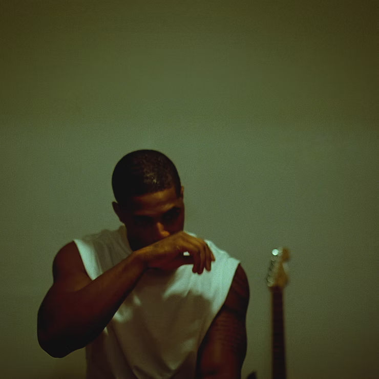 Man in a white sleeveless shirt wiping his face in a dimly lit room, with a guitar headstock in the background