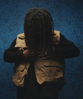 Person with long hair wearing a tan vest against a blue textured background, head bowed.