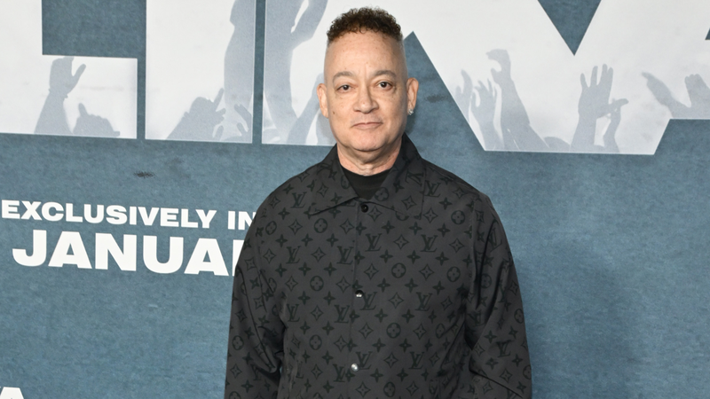 Man standing at a premiere backdrop, wearing a black patterned shirt with a slight smile.