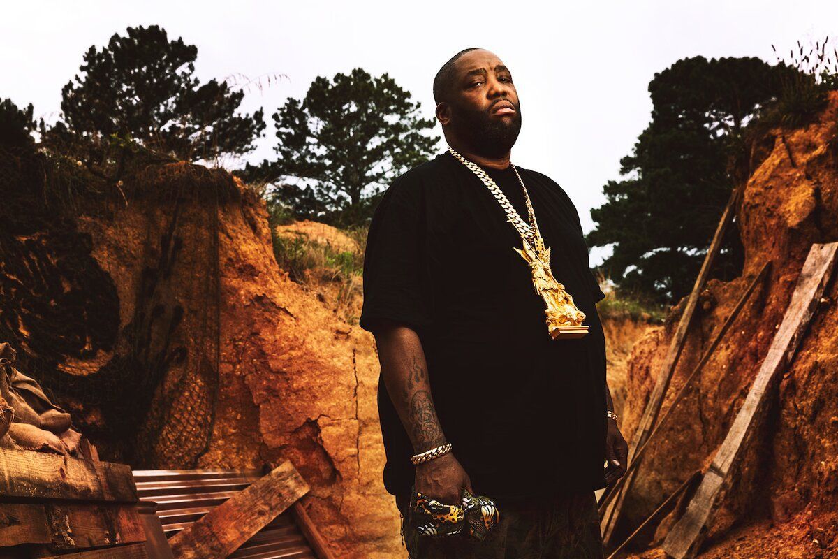 Man standing in a dirt excavation site between rocky walls, wearing a black shirt and gold chain.
