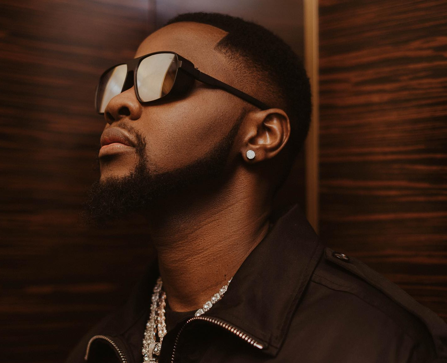 Man in dark sunglasses and black jacket against a warm wood-paneled background, looking upward.