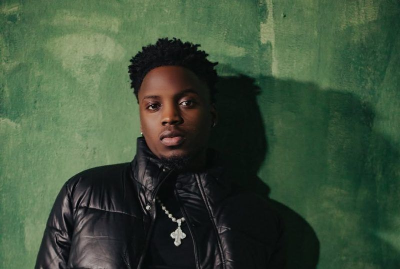 Man in a black jacket stands against a green wall, looking at the camera.