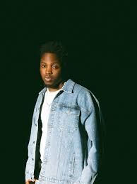 Person in a light denim jacket stands against a dark background.