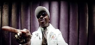 Man in white jacket and cap pointing toward the camera against a purple curtain backdrop