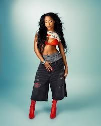 Standing in black wide-leg pants, red boots, and a cropped top against a teal background