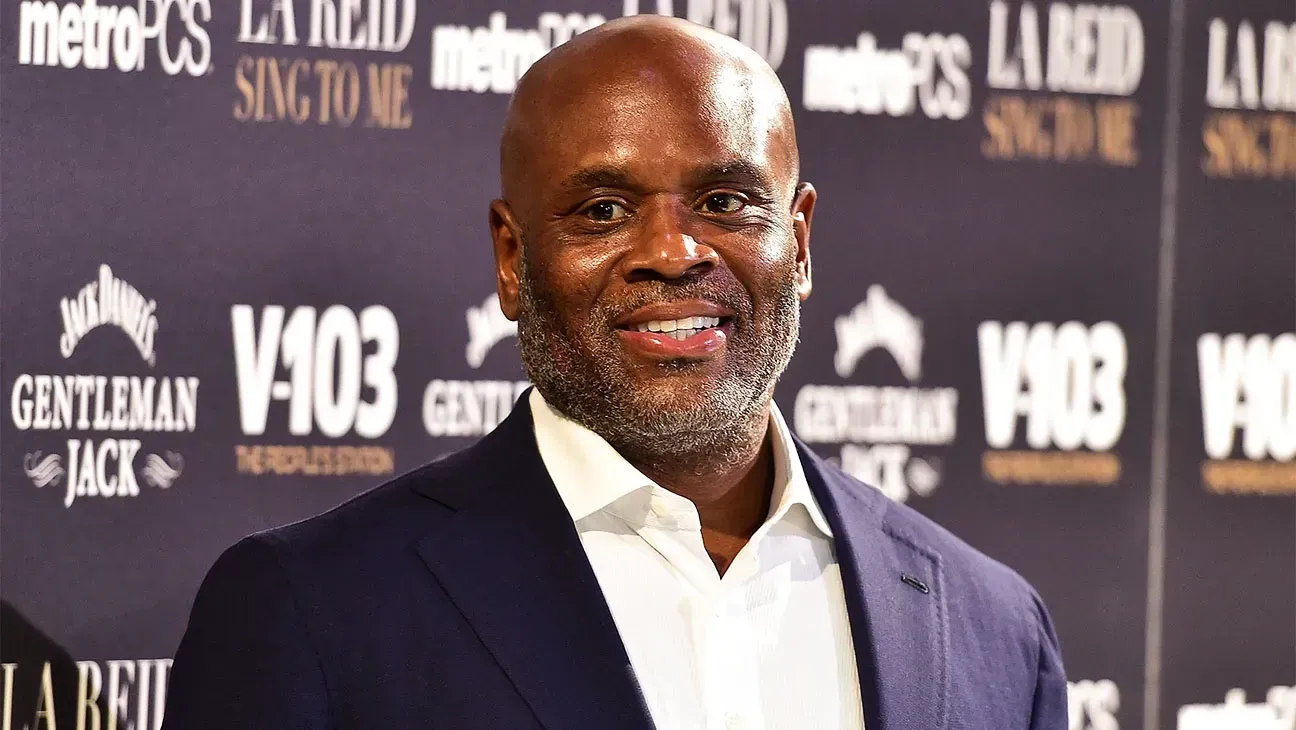 Man in dark suit and white shirt smiling at a red-carpet event with sponsor backdrop