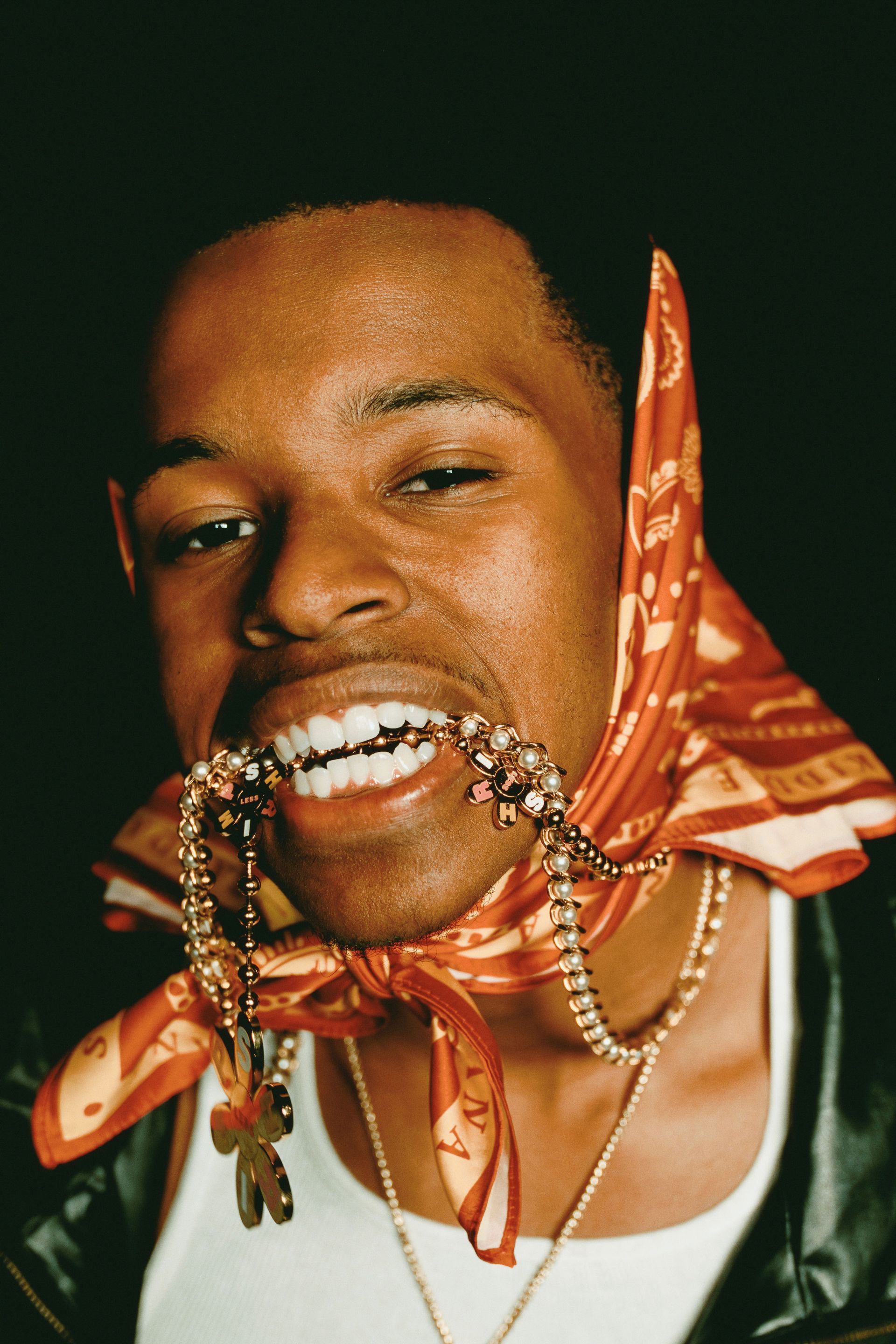 Person biting a beaded necklace, wearing a red bandana and black vest against a dark background