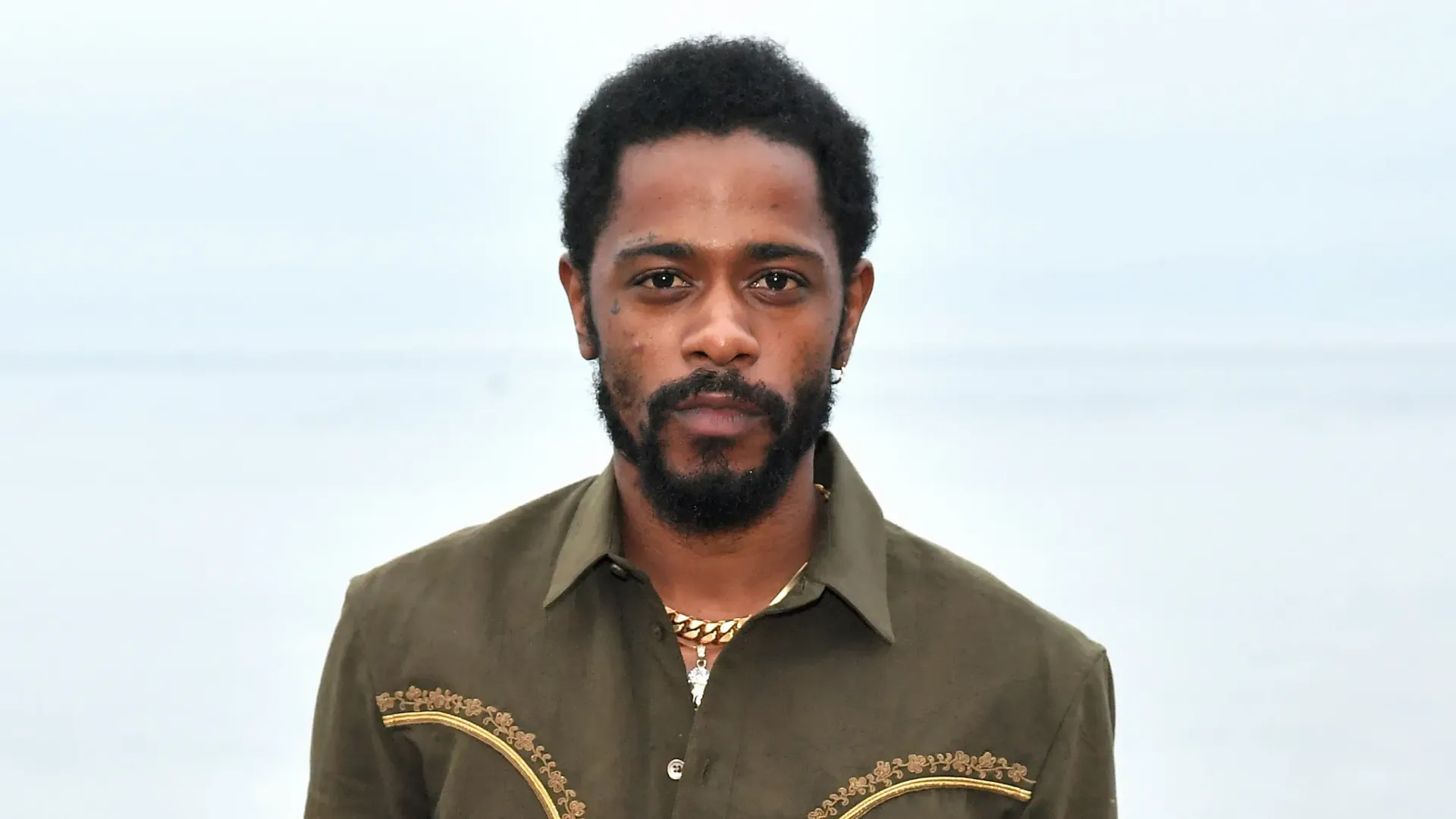 Portrait of a man with short curly hair and a beard, wearing an olive shirt against a pale background