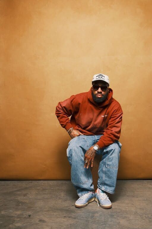 Man seated against an orange wall, wearing a rust sweatshirt, blue jeans, cap, and sunglasses