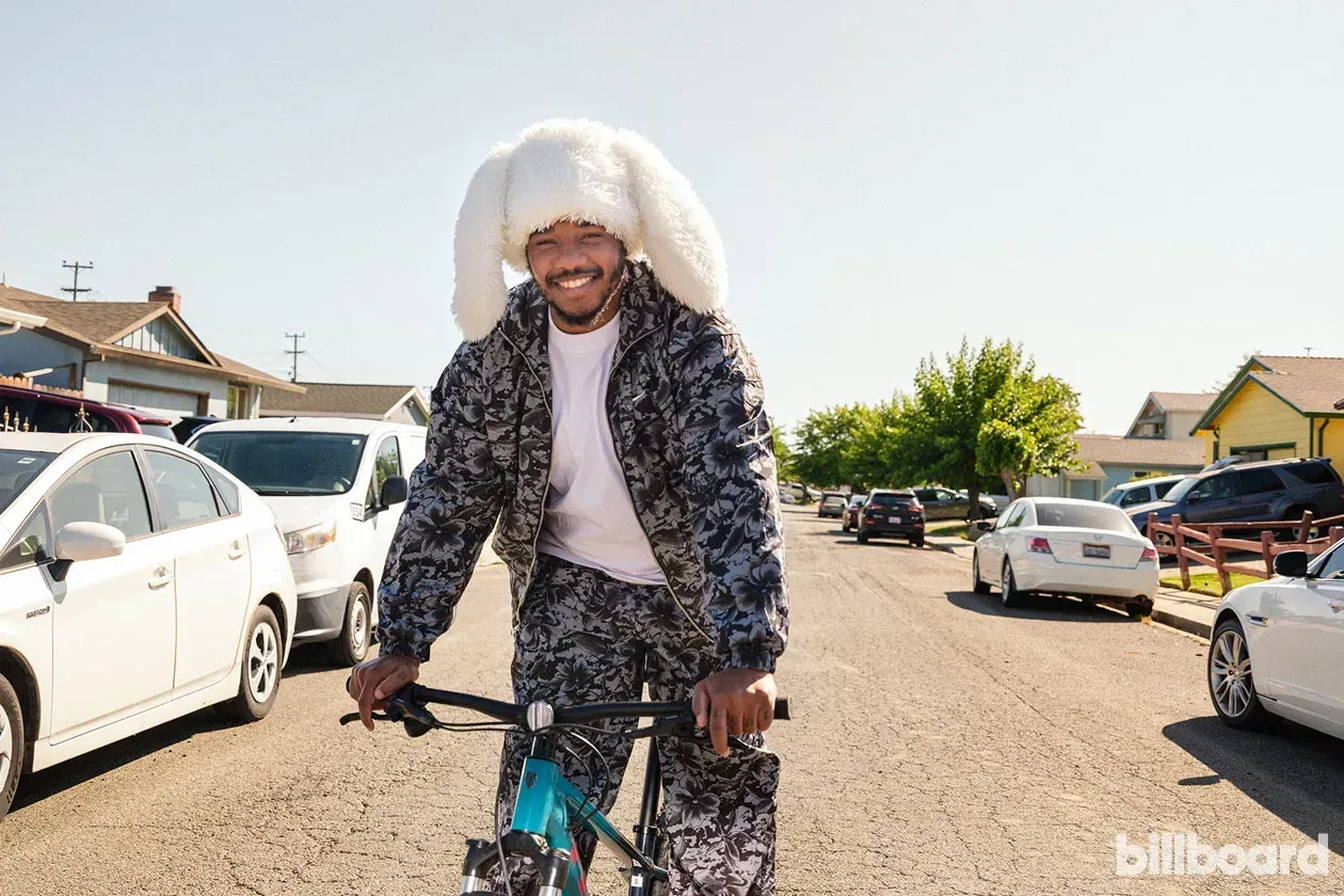 Person in a fur-trimmed hooded coat riding a bicycle on a sunny street with parked cars.
