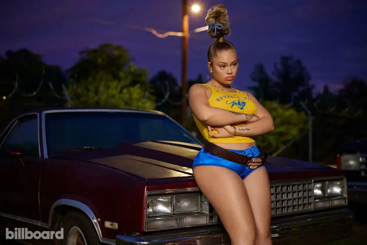 Woman posing in front of a burgundy classic car at night, arms crossed, with a Billboard logo in the corner