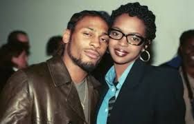 Two people posing together indoors, one in a brown jacket and the other in glasses and a dark blazer with a teal shirt.