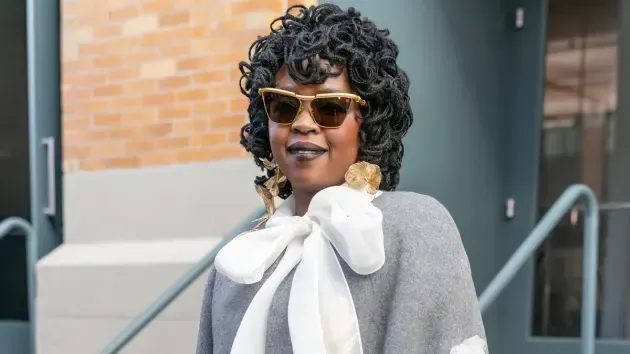 Person with curly hair and sunglasses wearing a gray top with a large white bow outdoors in front of a building.