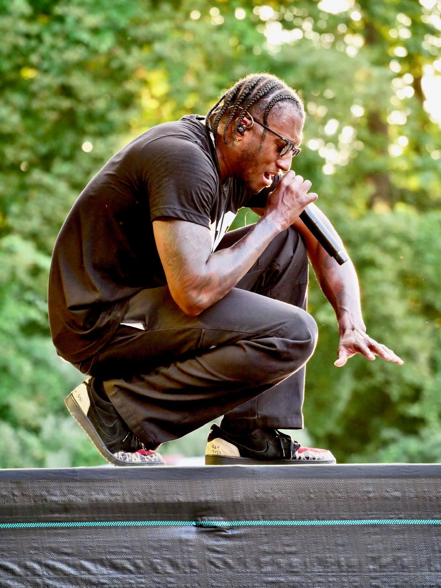 Man crouching onstage, holding a microphone and performing outdoors against a blurred green background