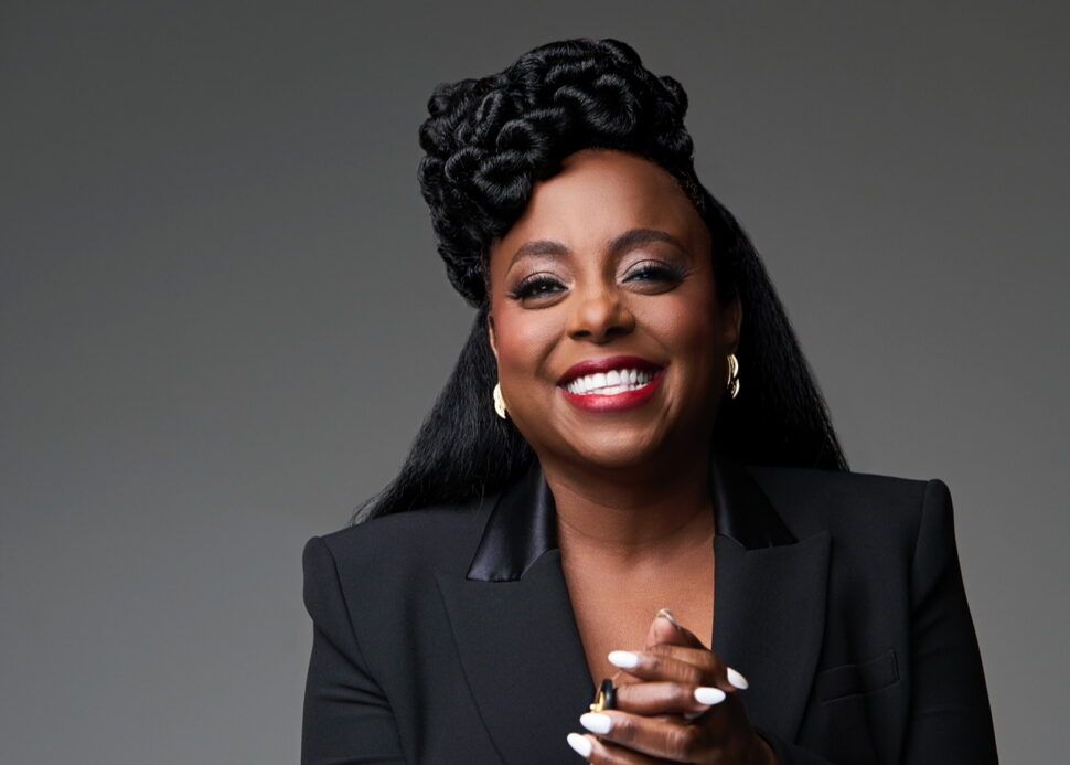 Smiling woman in a black blazer, hands clasped, against a gray background