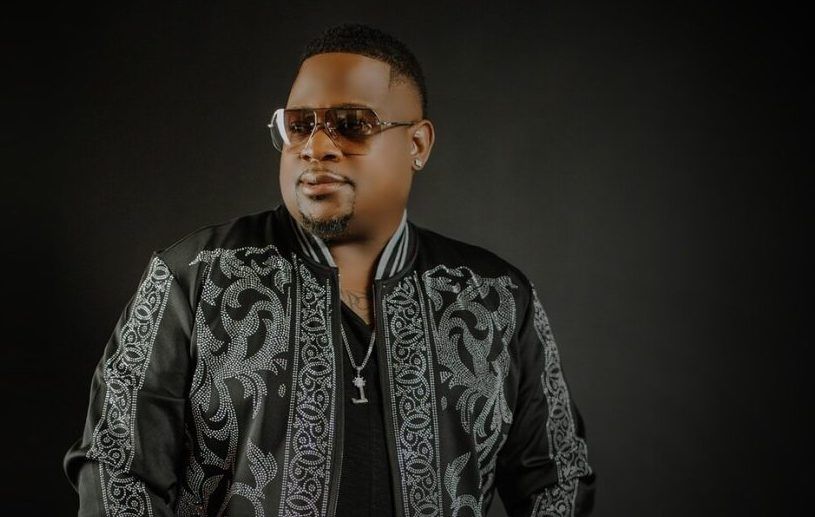 Man in sunglasses wearing a black patterned jacket against a dark background.