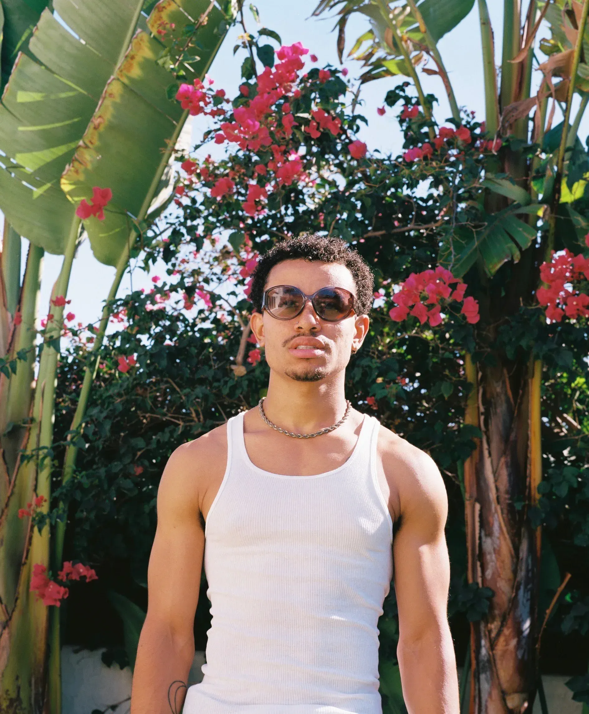 Man in sunglasses and white tank top standing outdoors among tropical plants and pink flowers.