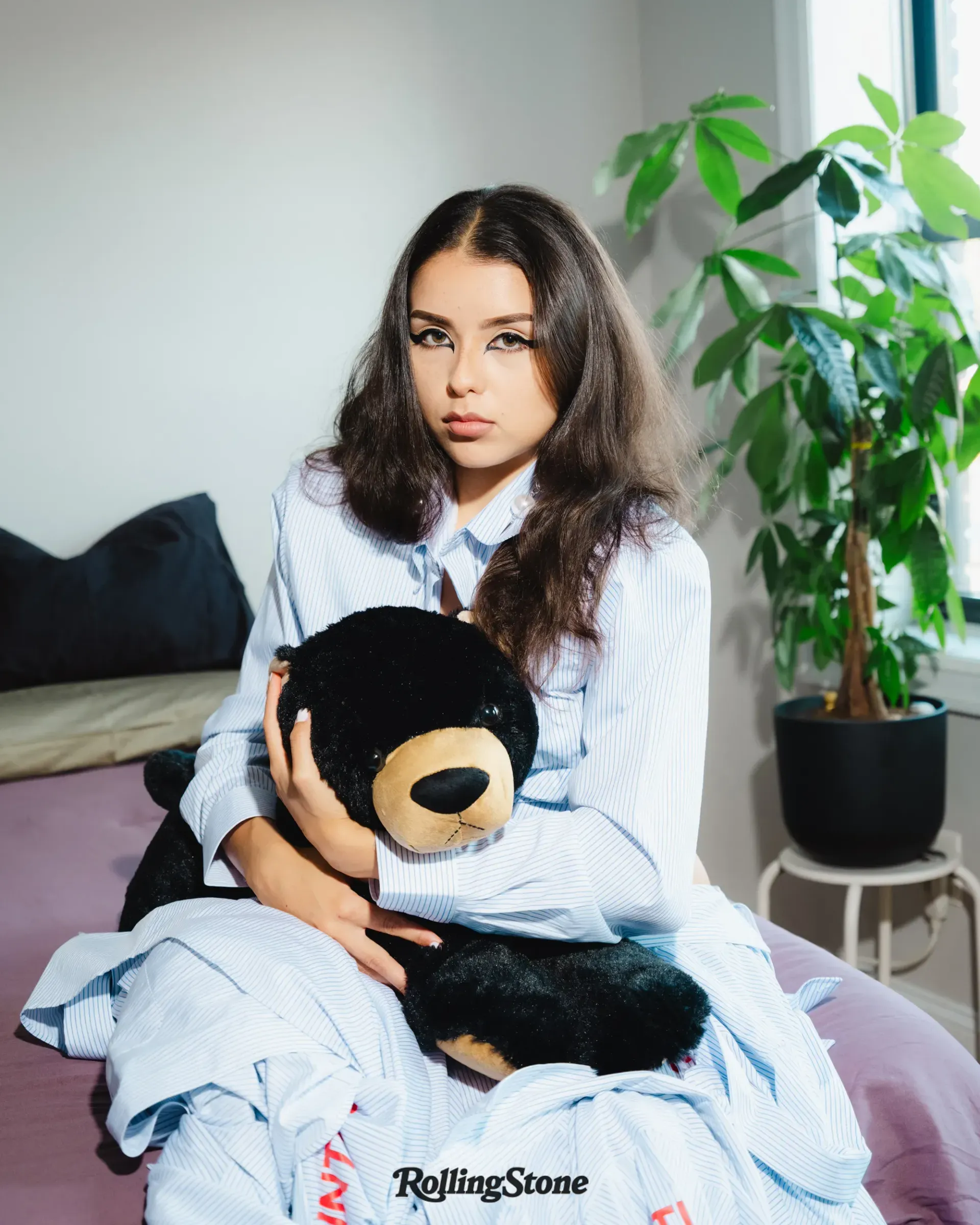 Woman sitting on a bed holding a black-and-tan teddy bear, with a potted plant beside her.
