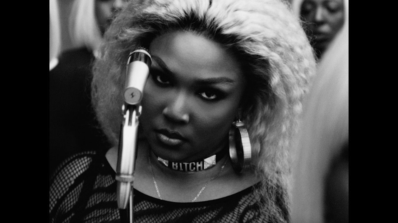 Close-up black-and-white portrait of a singer at a microphone, wearing a choker and fur hood, staring intensely