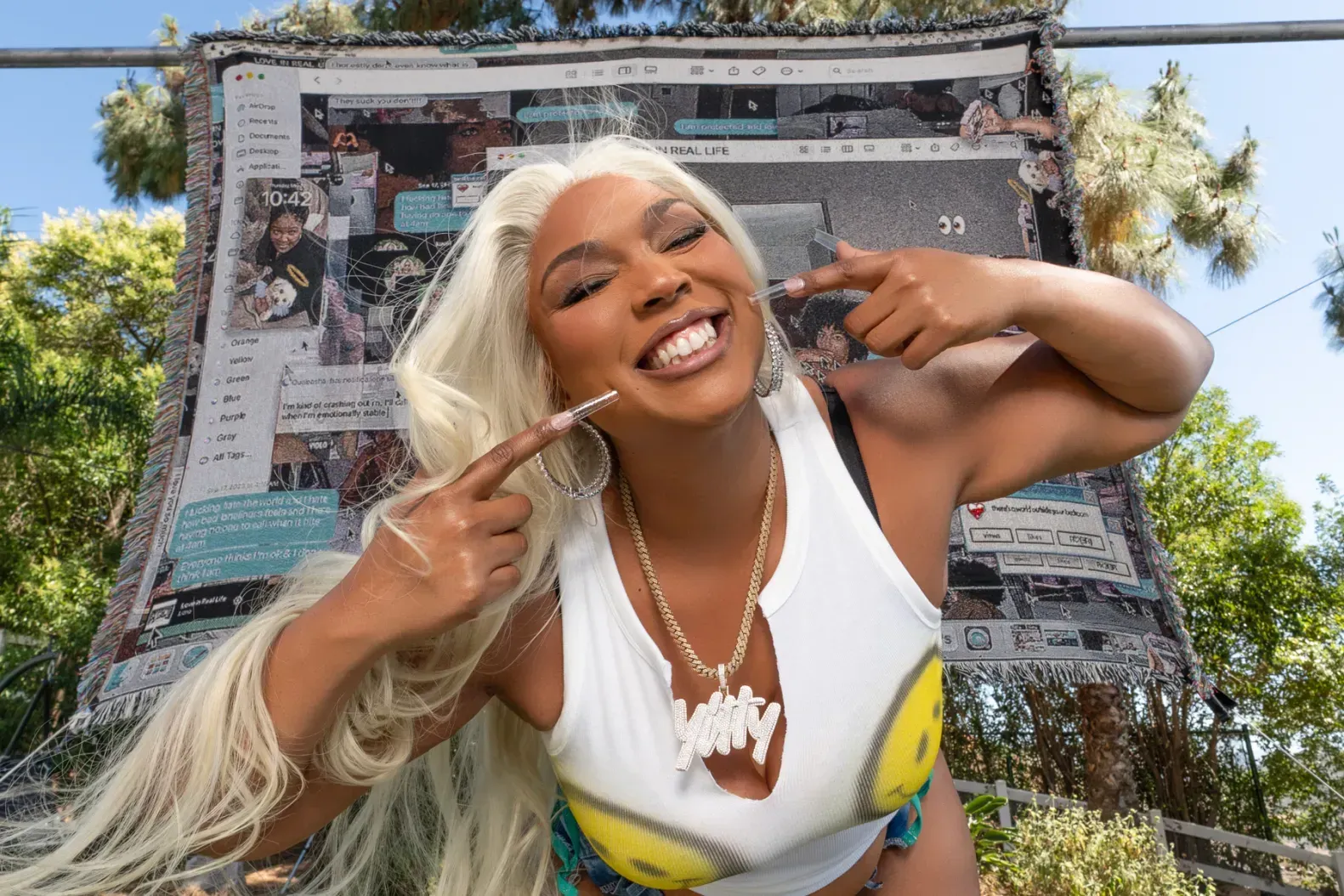 Smiling woman with long blonde hair posing outdoors in a white top, holding up a peace sign.