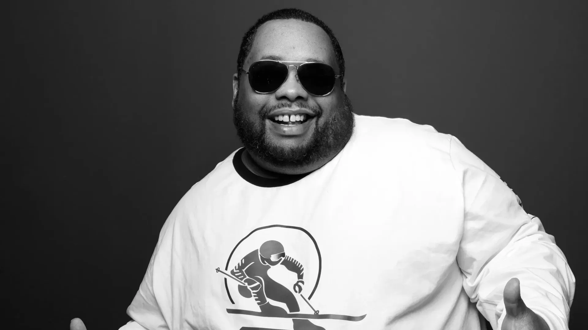 Smiling person in sunglasses and a white graphic sweatshirt against a dark background