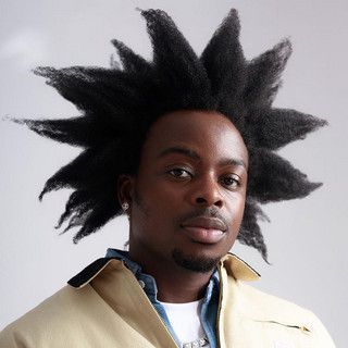 Portrait of a person with spiky black hair in a beige jacket against a light background