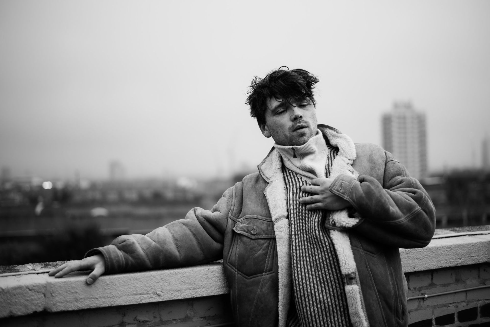 Person leaning on a rooftop wall, wearing a puffer jacket and scarf, with a city skyline behind.
