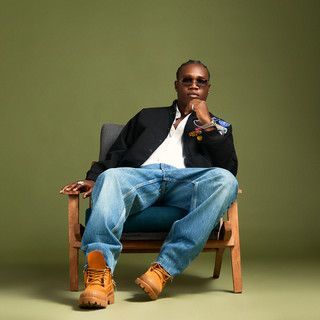 Person in sunglasses sits in a chair against a green wall, wearing jeans, black jacket, and tan boots.