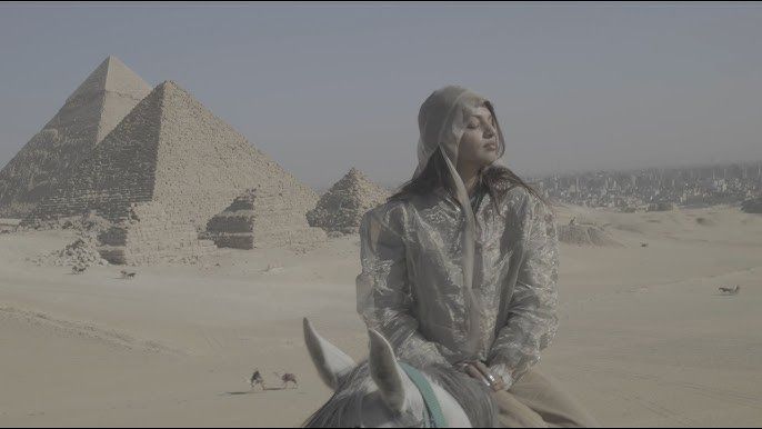 Person in silver jacket sitting in the desert with pyramids in the background, looking upward.
