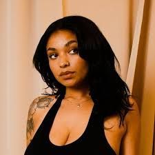 Woman in a black halter top, with dark hair and a shoulder tattoo, posing against a beige curtain.