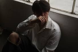 Person sitting on the floor by a window, wearing a white shirt and black ripped jeans, hand over face.