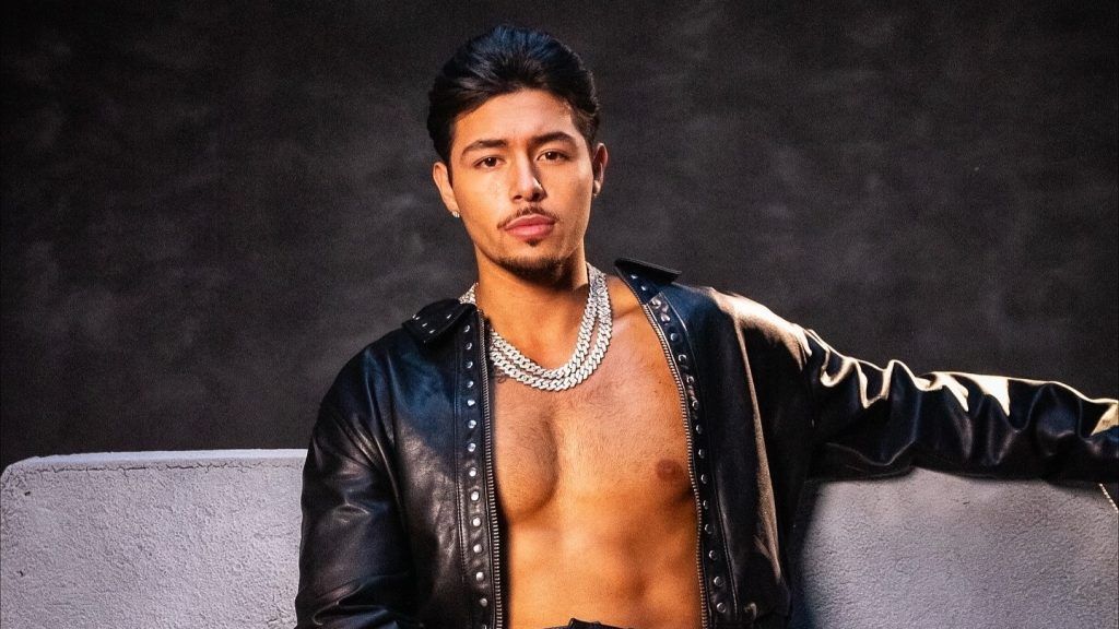 Portrait of a shirtless man in a black jacket and silver chain against a dark studio backdrop