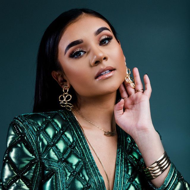 Portrait of a person in a green patterned outfit with gold jewelry, posing against a dark teal background