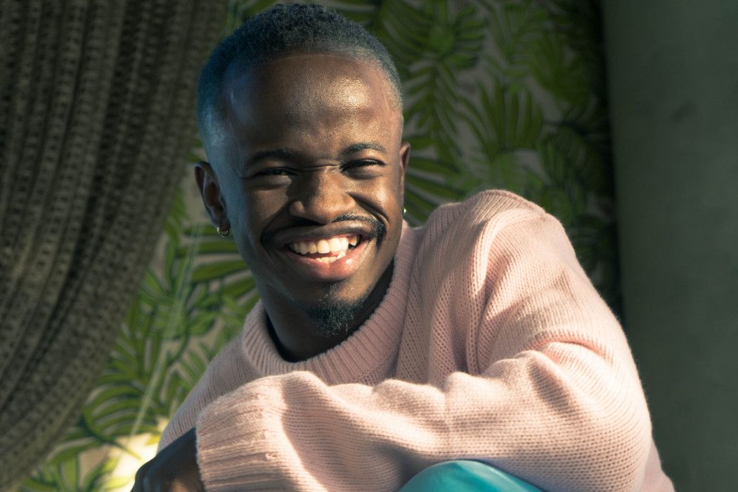 Smiling man in a light pink sweater seated by a leafy window