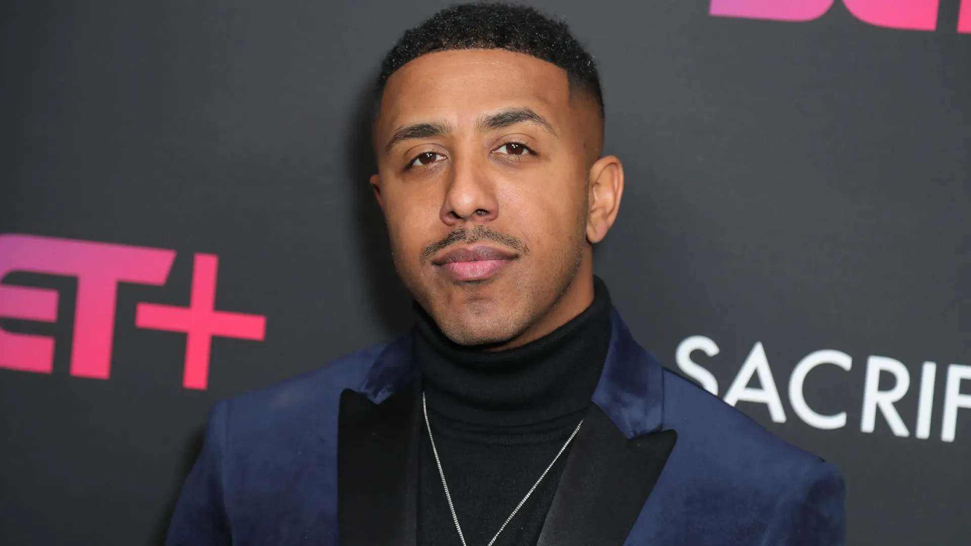 Man in a dark blazer and black turtleneck posing at a red carpet event with ET+ signage behind him