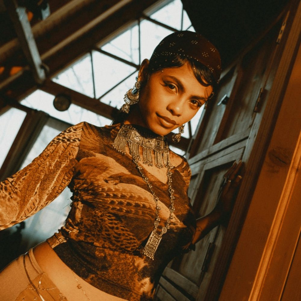Portrait of a person in a bronze lace top, leaning in a doorway with warm sunlight and wooden beams