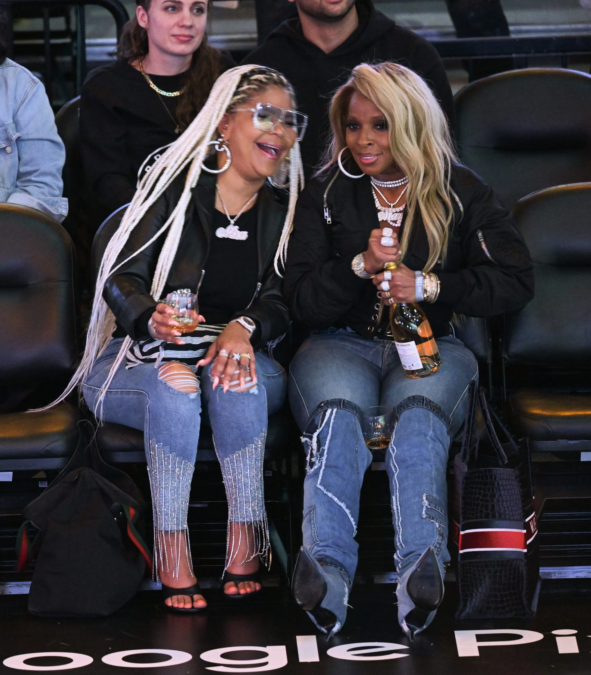Two women in black jackets and jeans sit courtside, smiling and holding drinks at a basketball game.