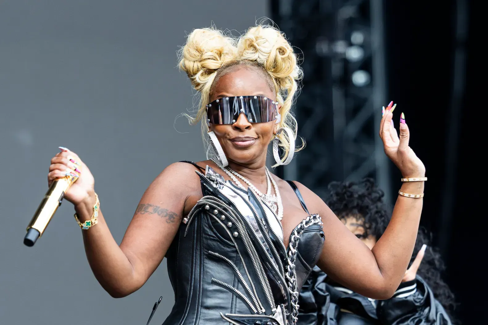 Singer onstage holding a microphone, wearing sunglasses and a black outfit, with both hands raised.