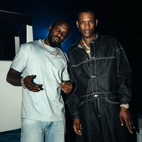 Two men posing indoors, one in a light T-shirt and one in a dark jacket, both wearing chains.