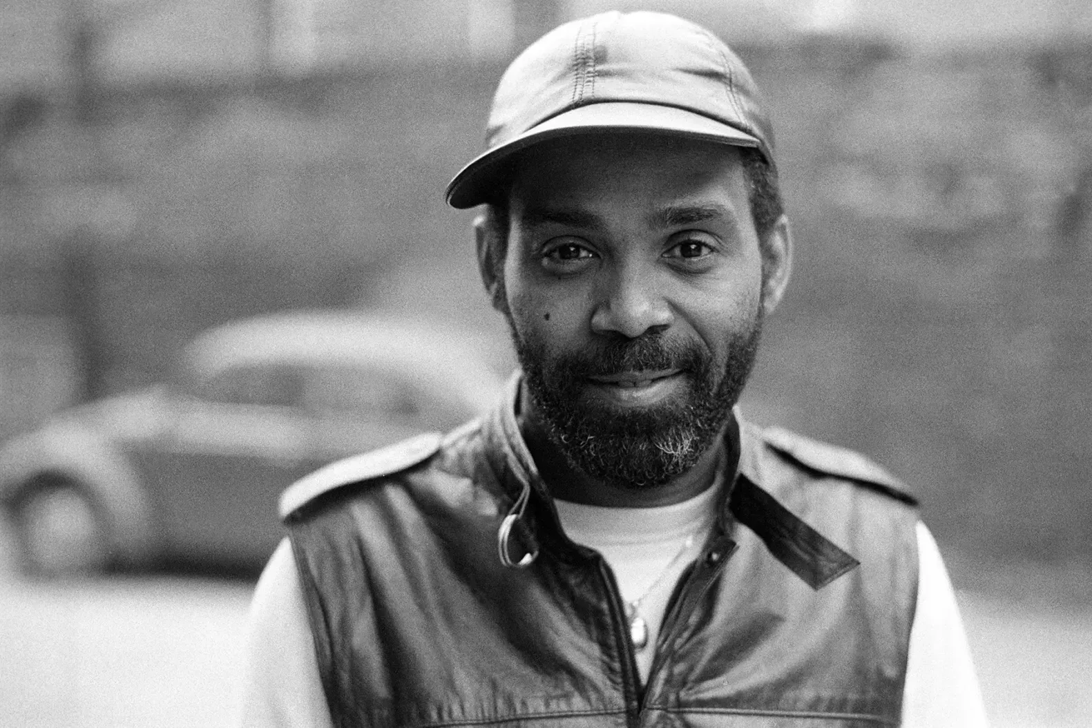 Black-and-white portrait of a man in a cap and vest, looking at the camera with a slight smile.