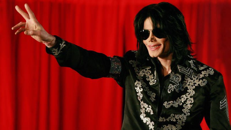 Michael Jackson in sunglasses and a black jacket, posing with a peace sign before a red curtain