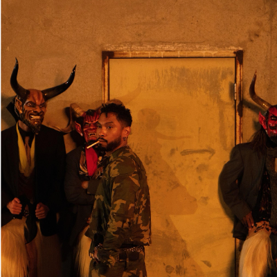People in horned masks stand against a yellow wall, one in camouflage in the foreground.
