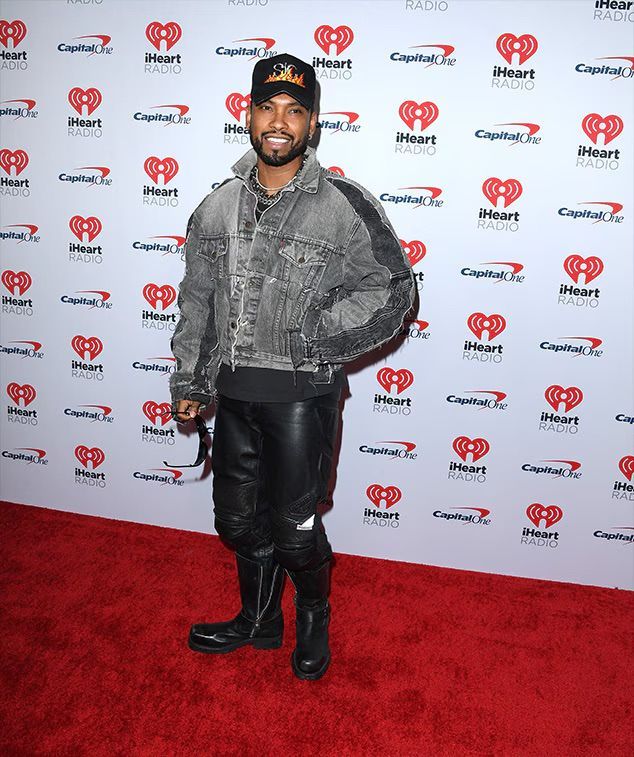A man in a black cap and gray jacket poses on a red carpet in front of an iHeartRadio backdrop.
