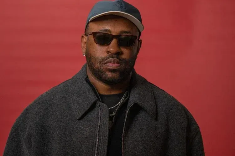 Man in sunglasses and cap wearing a dark coat against a red background
