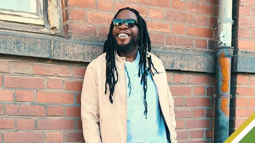 Man in sunglasses and beige jacket standing against a brick wall, smiling outdoors