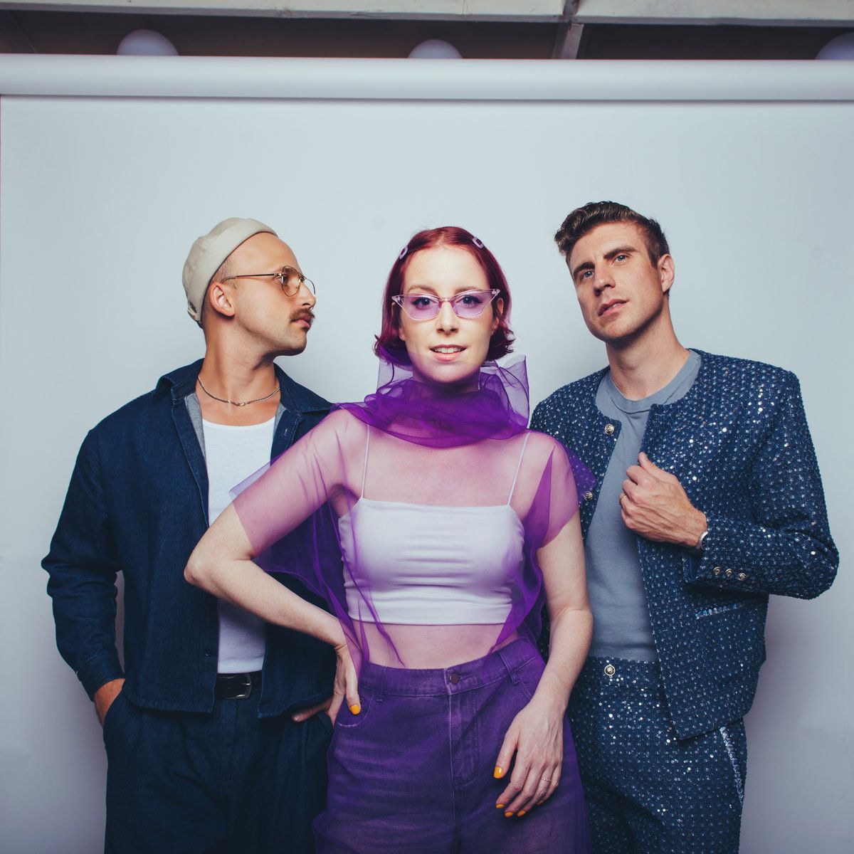 Three stylish people posing against a plain wall, with purple and blue outfits and sunglasses.
