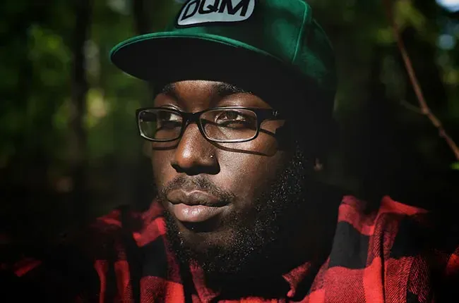 Man wearing glasses and a green cap, lit softly against a dark background, in a red plaid shirt