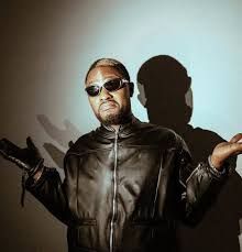 Man in black leather jacket and sunglasses, arms raised, against a plain wall with his shadow behind him